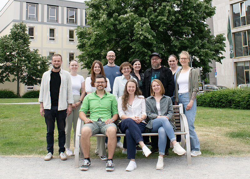 Gesamtes Team von INklusiv mit 12 Personen, einige sitzen auf einer Bank, andere stehen dahinter, im Hintergrund moderne Gebäude und Bäume